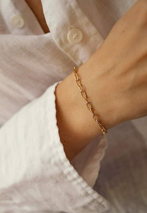 Pulsera de cadena de oro con eslabones ovalados y acentos texturizados, llevada en una muñeca, contrastando con una camisa de lino blanca.