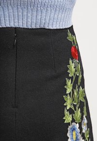 Falda de fieltro negra con vibrante bordado floral en rojo, azul y verde a lo largo de un lado. Suéter de punto gris mostrado en la parte superior.