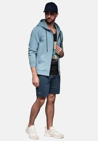 Jasnoniebieska bluza z kapturem na zamek, z przednią kieszenią, noszona na czarnym tank topie z grafiką, w połączeniu z ciemnymi szortami i białymi trampkami.