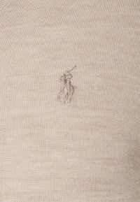 Beige stickad tröja med en subtil ribbad struktur och en liten broderad logotyp i matchande färg på vänster bröst.