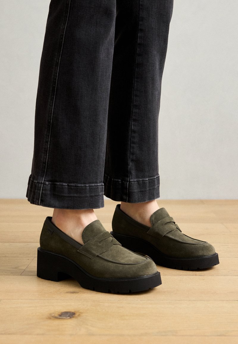 Mocassini in suede verde oliva con una spessa suola nera a piattaforma. Punta arrotondata, senza hardware visibile, abbinati a jeans neri larghi.