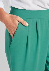 Grüne plissierte Hose mit glatter Textur und Seitentasche; der Bund ist flach und elastisch und sorgt für eine bequeme Passform.