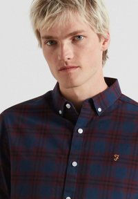 Camicia a scacchi con bottoni in blu navy e bordeaux, caratterizzata da un piccolo logo dorato, bottoni bianchi e un colletto classico. Tessuto morbido.