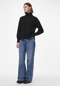 Črn pulover s turtleneck ovratnikom in rebrasto teksturo, v kombinaciji s širokimi modrimi jeans hlačami. Pulover ima spuščena ramena in sproščen kroj.