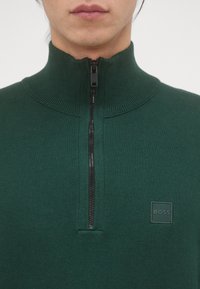 Donkergroene gebreide trui met een hoge geribbelde kraag, halve rits en een gestructureerde stof. Heeft een klein, vierkant logoetiket op de borst.