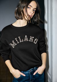 Sudadera negra con la palabra "MILANO" en letras de estampado de leopardo, cuello redondo y ajuste holgado, combinada con vaqueros de color azul claro.