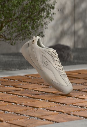 Light beige athletic shoe with mesh and suede materials, streamlined design, textured sole, and pronounced lacing system, hovering above wooden surface.