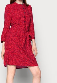 Robe florale rouge à manches longues, avec un devant à boutons et une ceinture à nouer. Présente un motif ludique et une coupe décontractée avec des fentes sur les côtés.
