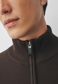Maglione marrone con zip e colletto alto a coste. Presenta una zip metallica con un tirante nero e un tessuto a maglia testurizzato, che mostra cuciture dettagliate.
