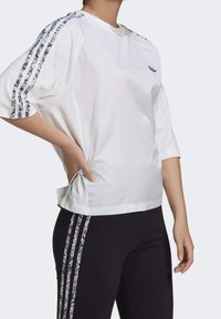 Vit sport-topp med raglanärmar och blommönstrade sidränder. Justerbar dragsko i midjan. Kombinerad med svarta leggings.