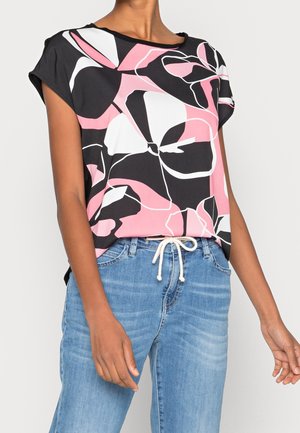Frau trägt ein schwarzes, pink-weißes Top mit abstraktem Blumenmuster kombiniert mit hellblauen Jeans, die mit einer weißen Kordel gebunden sind.