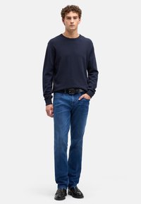 Morski sweter z okrągłym dekoltem w zestawieniu z niebieskimi jeansami. Model nosi czarne buty, trzyma ręce w kieszeniach. Materiał wydaje się miękki; jeansy są lekko postrzępione.