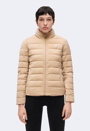 Vrouw met kort donker haar die een beige doorgestikt jack met een voorrits en zwarte broek draagt, staand tegen een effen achtergrond.