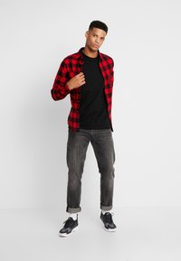 Camisa de franela a cuadros rojos y negros sobre una camiseta negra, combinada con jeans grises y deportivas negras, mostrando un estilo casual.