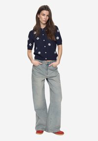 Tmavě modrý pletený cardigan s bílým květinovým vzorem, krátkými rukávy a zapínáním na knoflíky, v kombinaci s volnými světle modrými džínami a červenými botami.