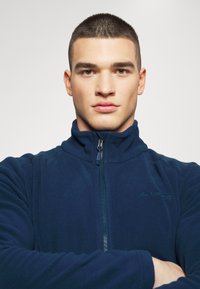 Tamsiai mėlynas flisinis džemperis su aukštu apykakle ir užtrauktuku. Funkcijos lygus paviršius ir minimalistinis ženklinimas kairėje pusėje.