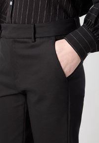 Pantalon noir sur mesure avec une texture lisse, doté d'une poche latérale et d'un devant plat. Porté avec une chemise noire à rayures.