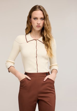 Frau mit langem, welligem Haar, trägt ein cremefarbenes langärmeliges Oberteil mit braunen Akzenten und hoch taillierte braune Hose, Hände in den Taschen, neutraler Hintergrund.