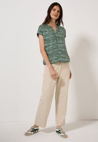 Blouse verte à manches courtes avec un motif zèbre, patte de boutonnage, associée à un pantalon large beige et des baskets grises avec des accents verts.