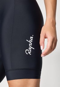 Mustat pyöräilyshortsit, joissa on valkoinen "Rapha." logo. Liukas, joustava kangas, korkea vyötärö ja kontrastiväriset ompeleet helman yli.
