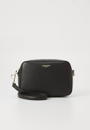 Black textured leather rectangular crossbody bag with gold zipper and "Lacoste Paris" logo on front, detachable strap coiled below.