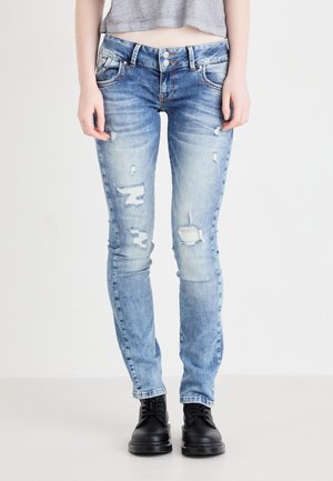 Person trägt hellblaue, zerrissene Skinny-Jeans und schwarze Schnürstiefel und steht vor einem schlichten weißen Hintergrund.