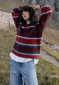 Gestreifter Wollpullover in Rot und Blau mit lockerem Schnitt, getragen über einem weißen Hemd, kombiniert mit hellblauen Jeans in einer natürlichen Outdoor-Umgebung.
