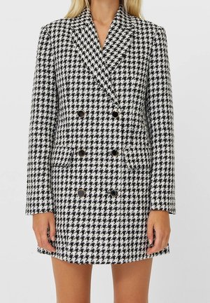 Woman wearing a black and white houndstooth patterned double-breasted blazer dress with black buttons and long sleeves.