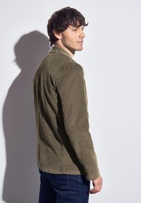 Chaqueta de pana verde oliva con un corte entallado, diseño de un solo botón y detalles de botones en las mangas, combinada con jeans azul oscuro.