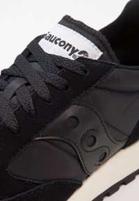 Black athletic shoe with a mix of suede and synthetic materials. Features a textured pattern and prominent Saucony branding on the tongue.