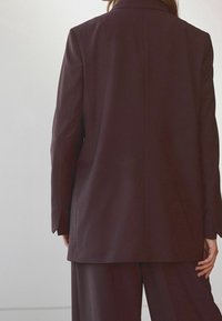 Blazer bordeaux avec une coupe ajustée, des revers crantés, une fente simple au dos et des manches droites. Texture lisse et coupe structurée.