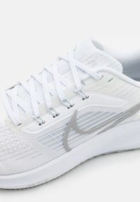 Zapatilla deportiva blanca con parte superior texturizada, detalles en plateado y cordones blancos. Cuenta con una suela acolchada con marcas visibles y un diseño ergonómico.