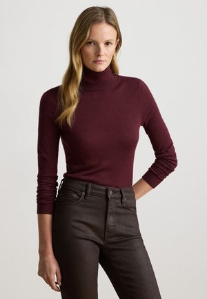 Frau trägt einen eng anliegenden burgunderfarbenen Rollkragenpullover und hoch taillierte schwarze Jeans, steht mit einer Hand auf der Hüfte vor einem schlichten Hintergrund.