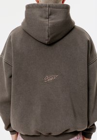 Sweat à capuche gris surdimensionné avec une poche kangourou avant, un tissu texturé et un logo brodé en marron clair au dos. Conception à capuche.