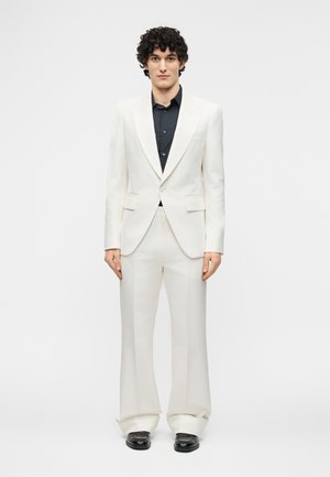 Νεαρός άνδρας με σγουρά μαλλιά, φοράει λευκό κοστούμι, μαύρο πουκάμισο και μαύρα παπούτσια, στέκεται με το πρόσωπο προς τα εμπρός μπροστά σε απλό γκρι φόντο.