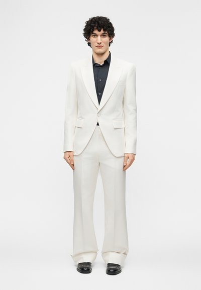 Jeune homme aux cheveux bouclés portant un costume blanc, une chemise noire et des chaussures noires, debout face à l'avant sur un fond gris uni.