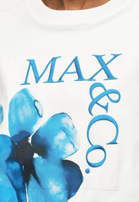 Λευκό πουκάμισο με μεγάλο μπλε λουλουδάτο σχέδιο και μπλε κείμενο "MAX & Co." τυπωμένο μπροστά.