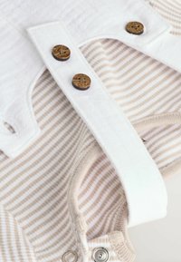 Baumwoll-Babykleidung mit weißen Trägern und Holzknöpfen sowie einem beige-weißen Streifenmuster. Mit Druckknopfverschluss für ein einfaches Ankleiden.