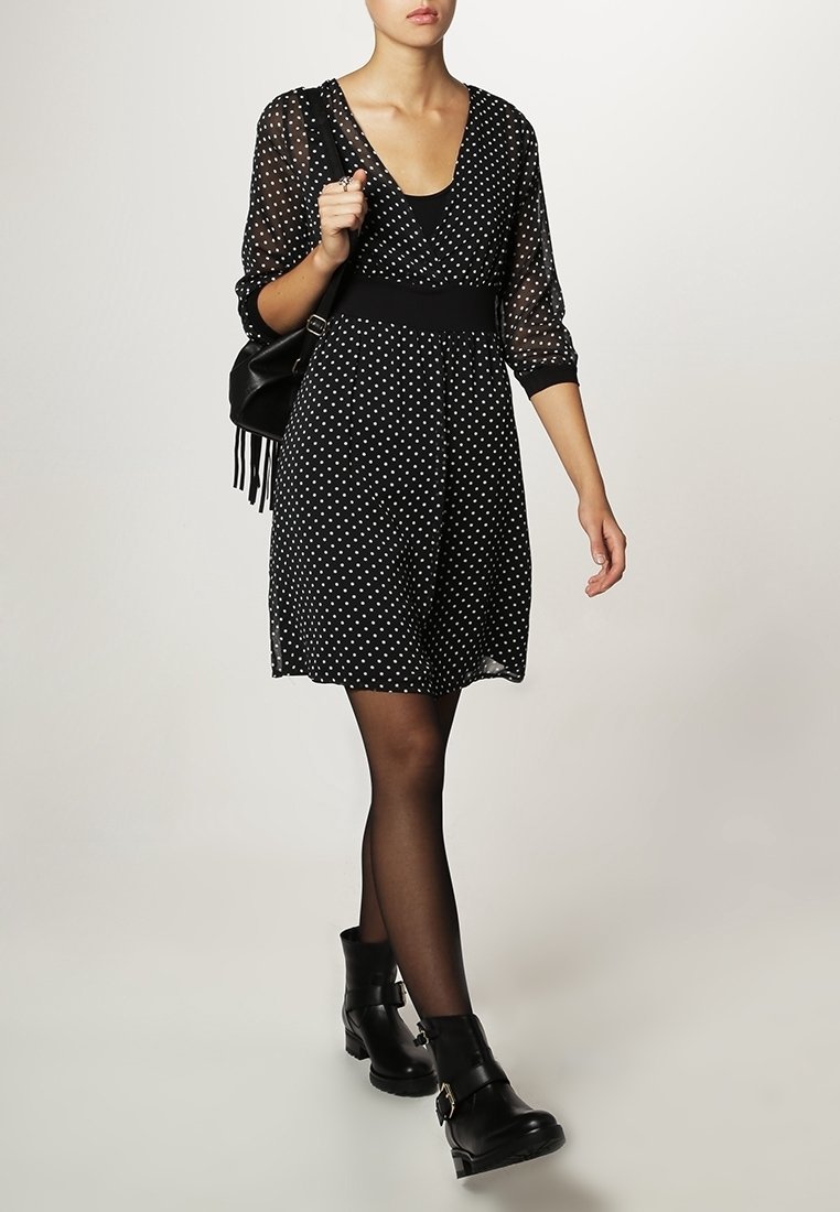 Femme portant une robe noire à pois blancs, manches transparentes, collants noirs, bottines noires et un sac à épaule noir.