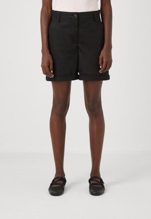 Persona que lleva pantalones cortos negros con puños, una camiseta de color claro y zapatos estilo Mary Jane negros, de pie contra un fondo liso y claro.