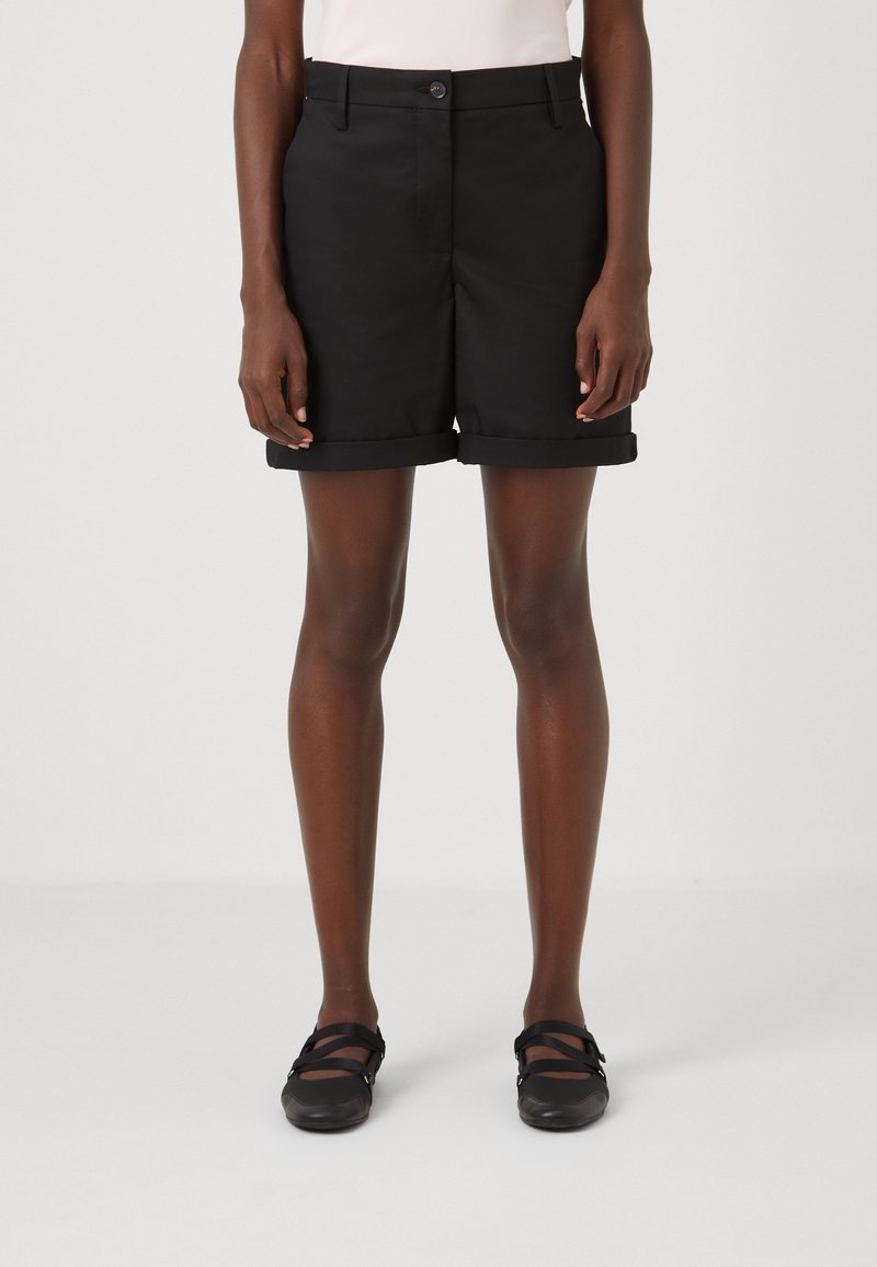 Personne portant un short noir à revers, un haut clair et des chaussures noires de style Mary Jane, se tenant devant un fond clair uni.