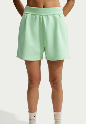Personne portant un short vert clair à taille élastique avec poches latérales et chaussettes blanches Nike, debout devant un fond uni.