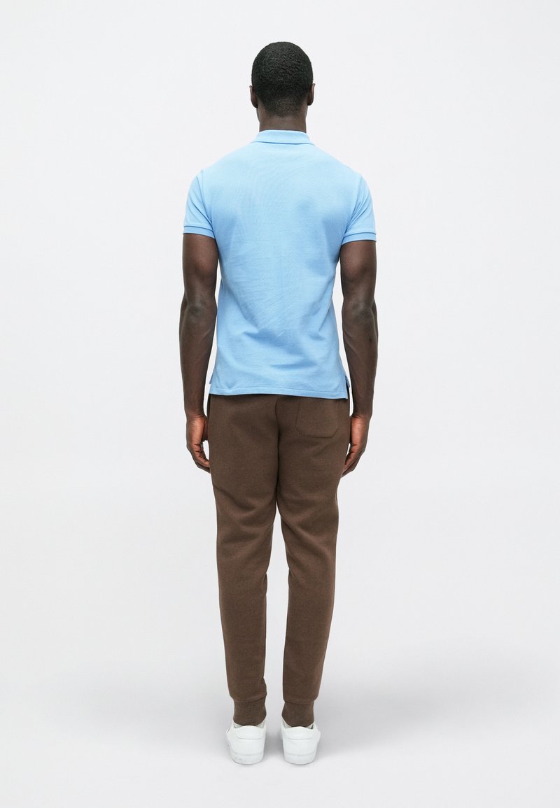 Blauwe polo met korte mouwen, gecombineerd met een bruine joggingsbroek en witte sneakers, gezien vanaf de achterkant tegen een effen achtergrond.