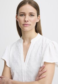 Blusa bianca con maniche a palloncino corte, caratterizzata da un motivo a pois testurizzati, collo alla mandarino e una sottile pattina sul davanti.