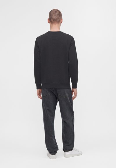 Homme portant une chemise noire à manches longues, un jean foncé et des baskets blanches, debout de dos sur un fond blanc uni.