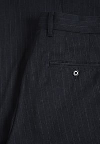 Pantalon bleu marine foncé à rayures fines, avec une ceinture passant dans des boucles, une poche arrière avec fermeture à bouton, et un tissu texturé.