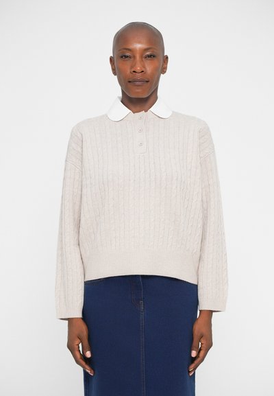 Pull en tricot beige avec un col blanc, doté d'une fermeture à boutons à l'avant et d'une coupe décontractée. Porté par-dessus une jupe marine.