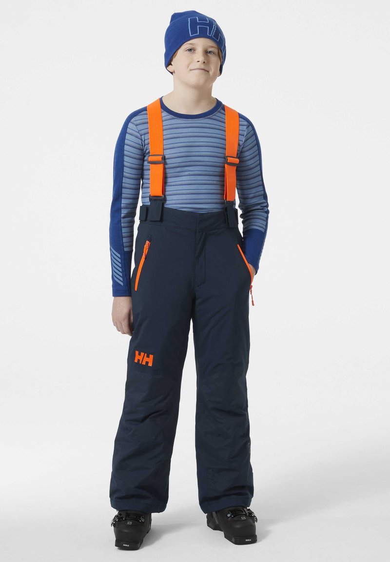 Pantalones de nieve en color navy con tirantes naranjas y cremalleras, combinados con una camiseta de manga larga a rayas azules y un gorro azul con un logo.