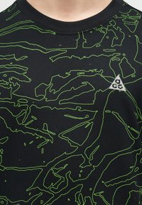 Μαύρο πουκάμισο με αφηρημένα σχέδια σε σχήμα γραμμών νέον πράσινου και μικρό λευκό τριγωνικό λογότυπο με την επιγραφή "acg" στο στήθος κοντά στη λαιμόκοψη.