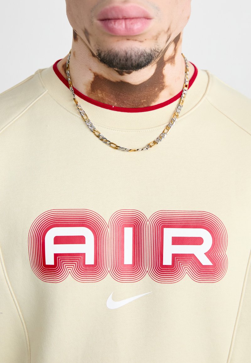 Beige Sweatshirt mit auffälligem roten "AIR"-Grafik, geripptem Kragen und weißem Nike-Logo. Eine silberne und goldene Kettenkette ist sichtbar.
